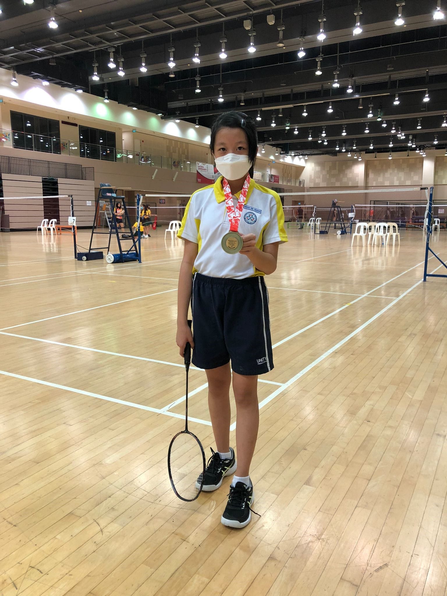 A young girl in a white and yellow polo shirt holds a badminton racket and displays a gold medal in an indoor court.